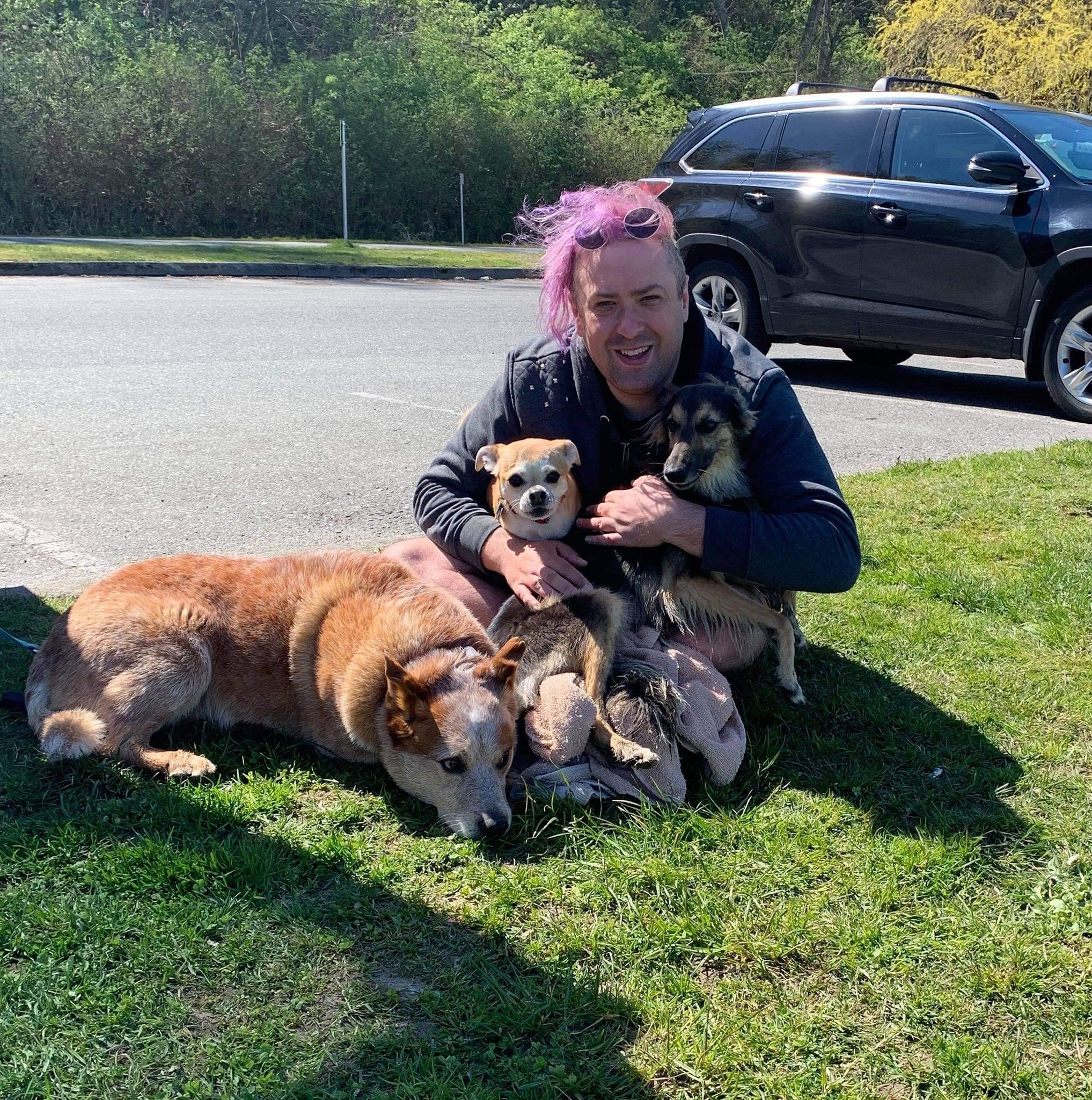 Lucian, co-owner of Douglas Fur Pets, sitting on the grass with three happy dogs during a Vancouver dog walk, showing the care and love behind our professional group dog walking service.